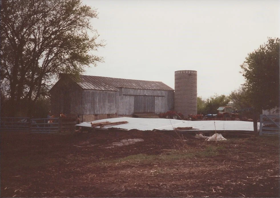 A farm scene with a tall barn, a silo, and several trucks and cars parked nearby. A large white tarp covers a section of the ground.