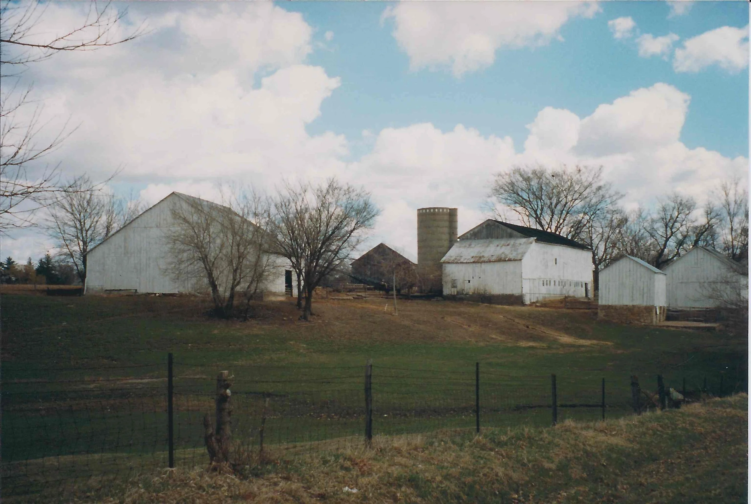 A rural farm scene with white barns, leafless trees, a metal silo, and a grassy area, under a partly cloudy sky.