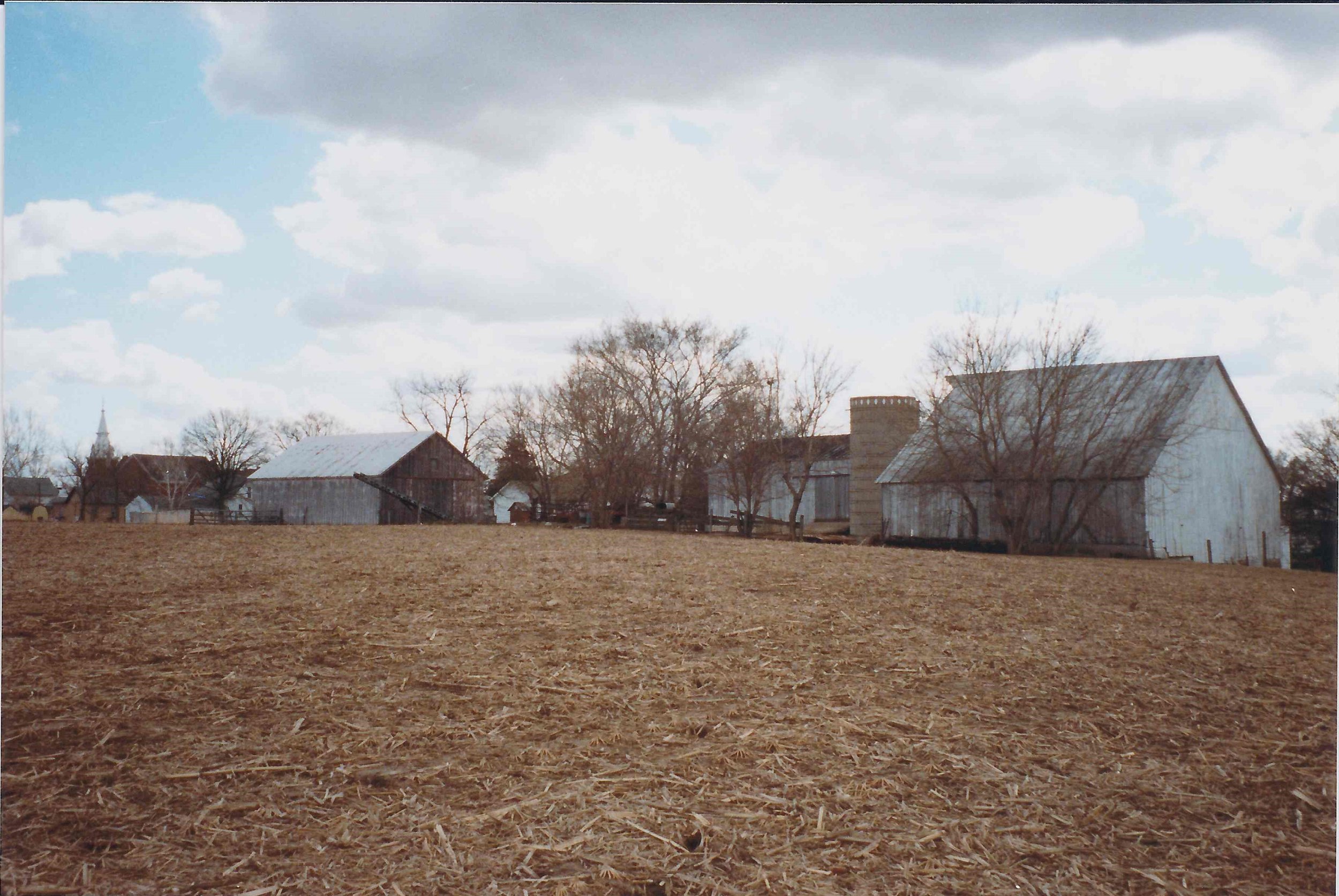 A rural farmland scene with several weathered barns and trees, under a partly cloudy sky.