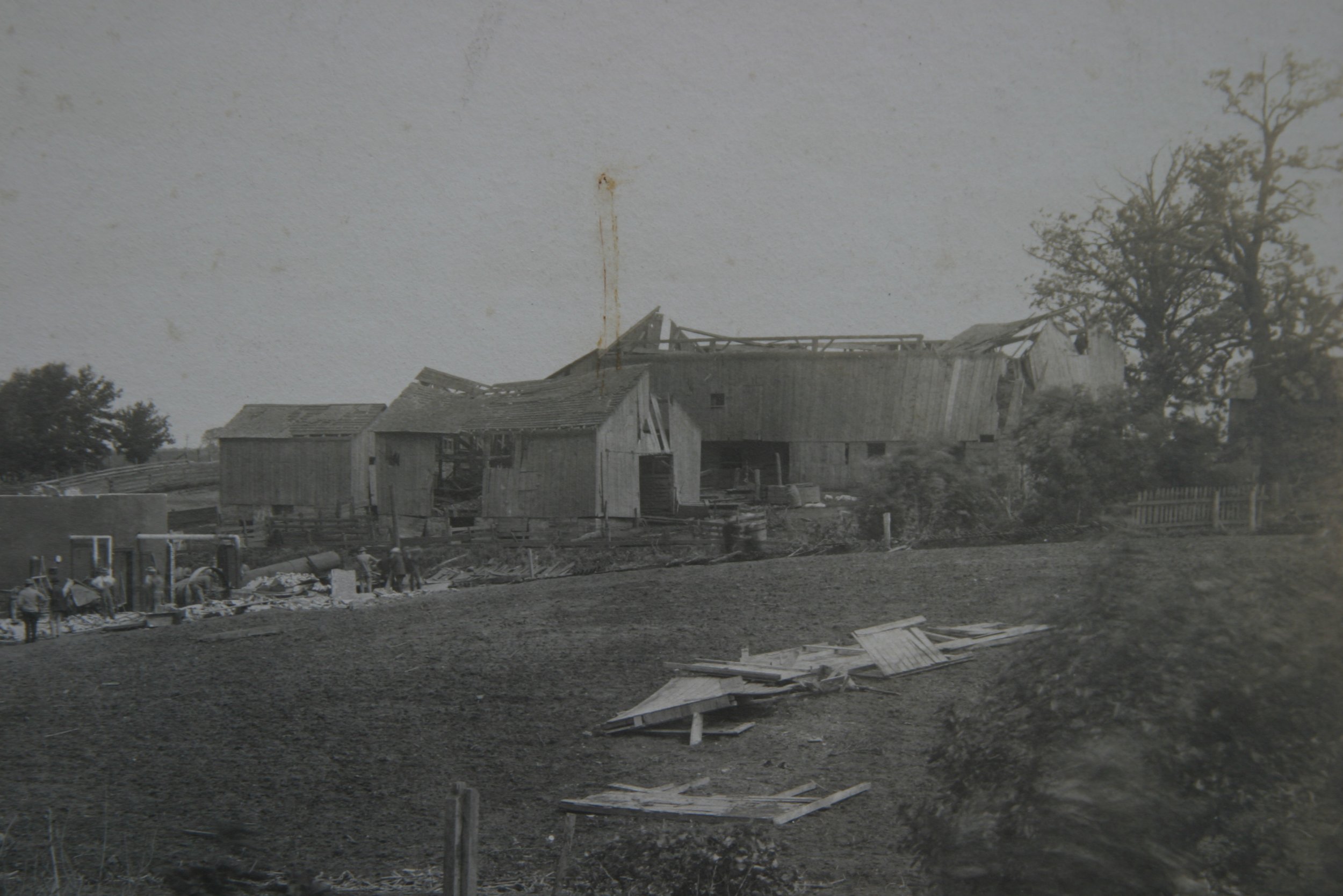 A black and white photo showing a partially destroyed barn with damaged walls and roof, debris scattered in the foreground, and trees in the background.