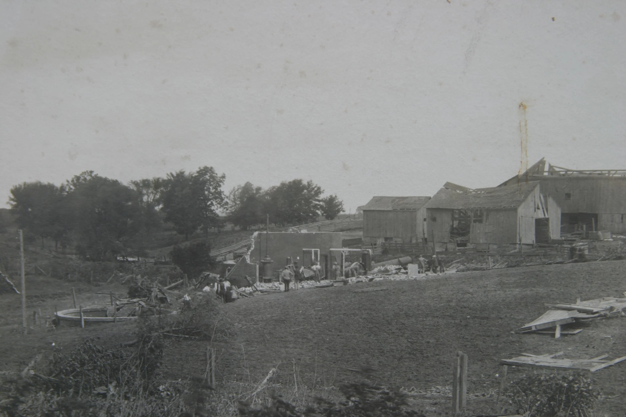 Black and white photo of a rural area with damaged buildings, construction activity, and some trees in the background.