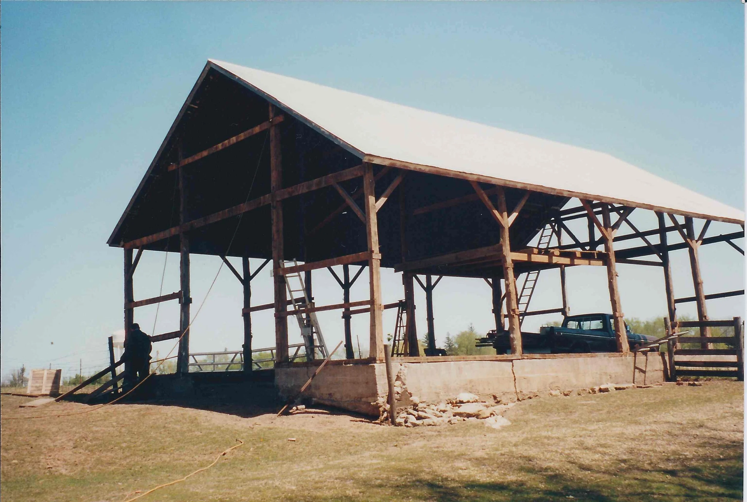 Wooden structure under construction with a white roof, ladder inside, and a person working on the left side, outdoors on grassy ground.