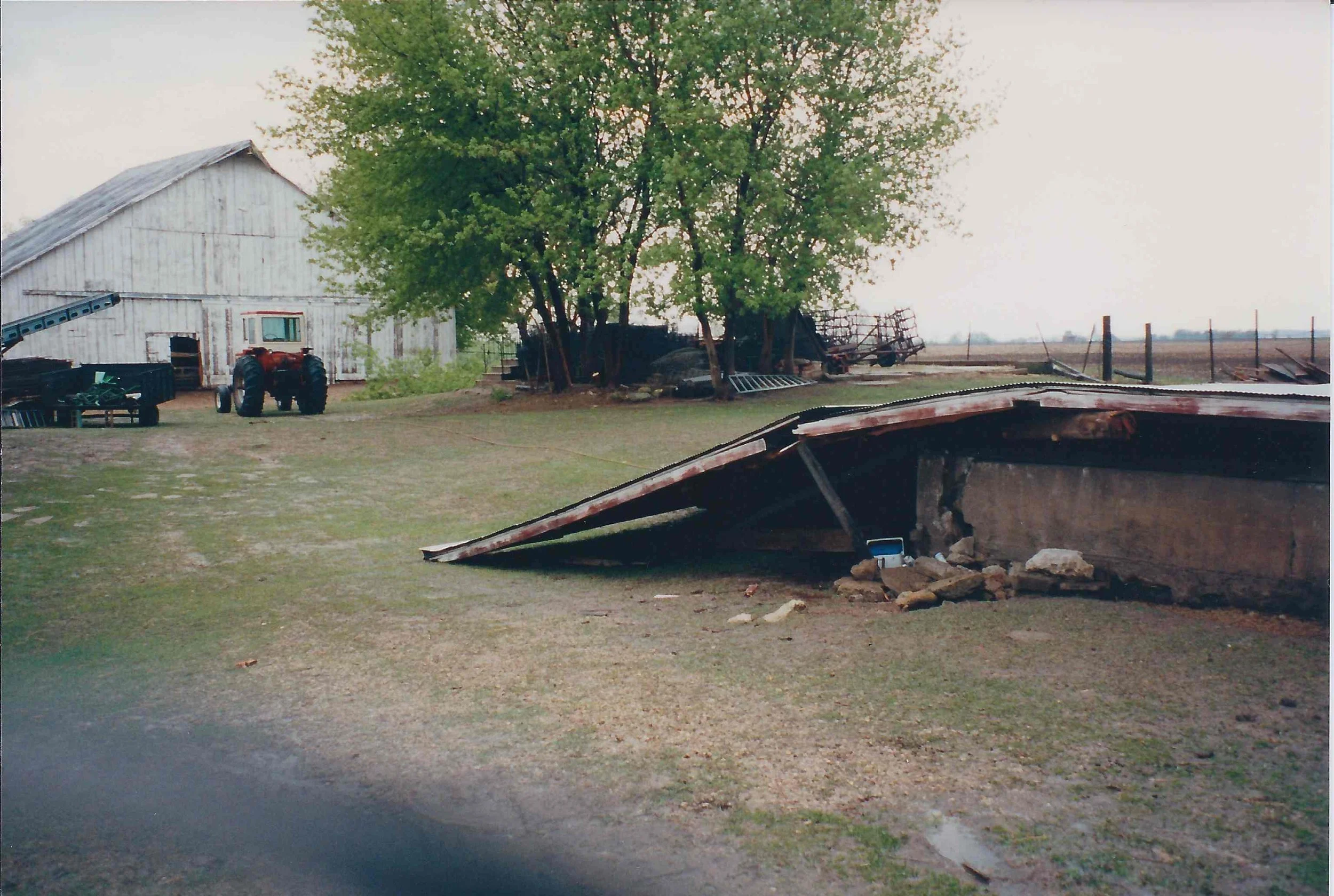 A rural farm scene showing a damaged barn roof, a tractor, and farm equipment beside trees and a white wooden building.