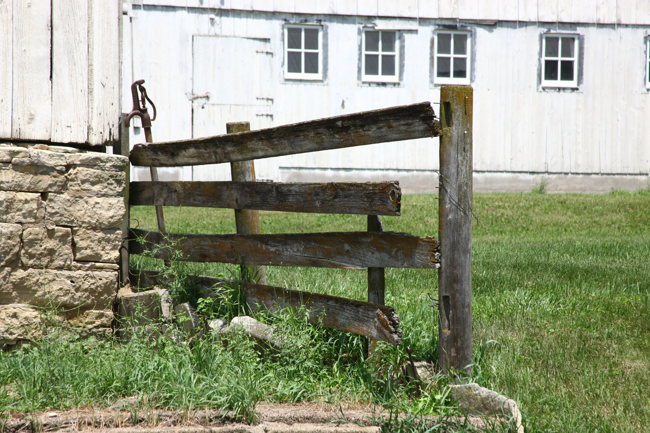 A weathered wooden fence next to a stone pillar, with an old metal tool hanging on the stone, in front of a white wooden building with multiple windows.
