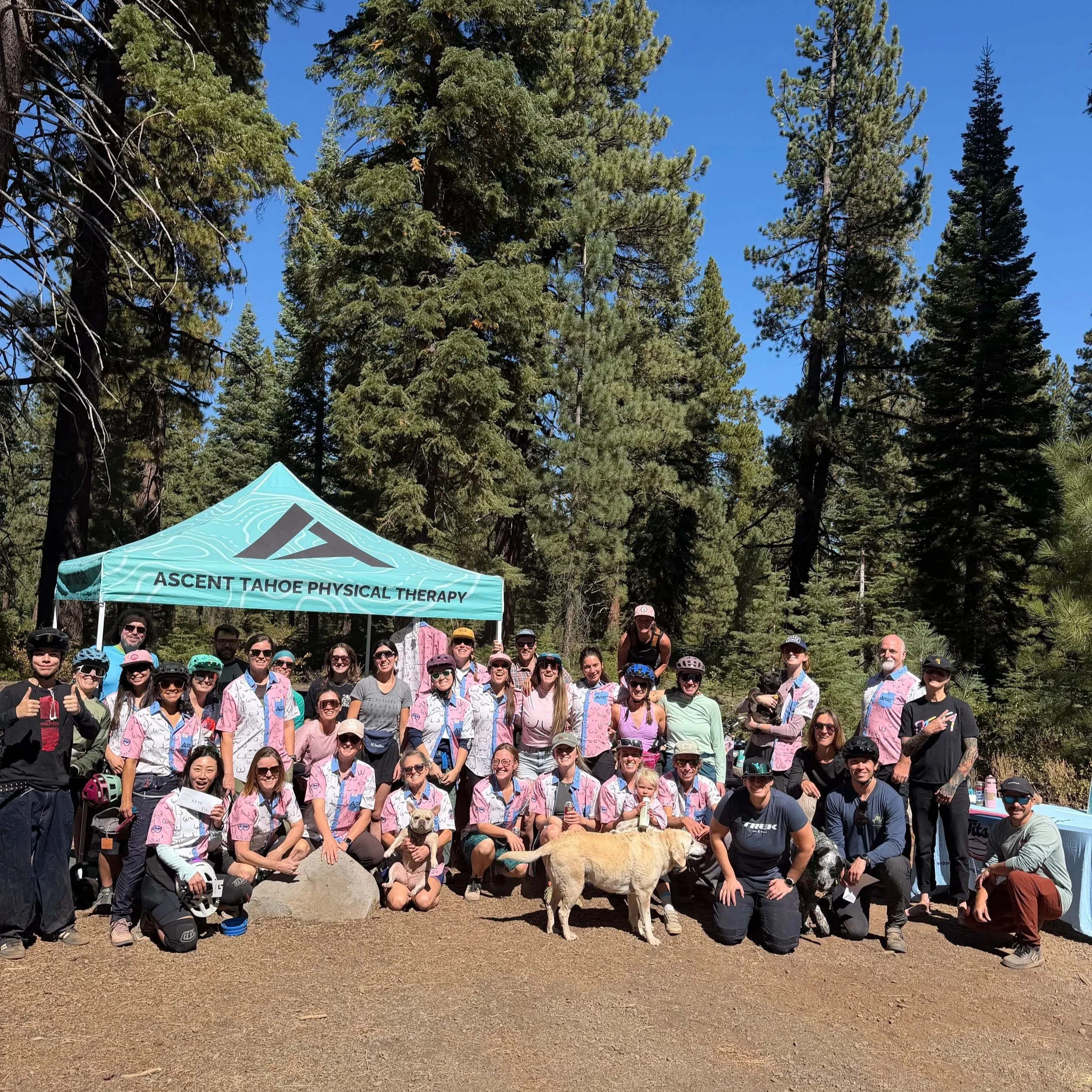 Group of people gathered outdoors in front of tall pine trees with a blue sky, wearing colorful outdoor clothing and helmets, some wearing sunglasses, posing for a group photo with a dog in front, and a teal tent with black text that reads "Ascent Tahoe Physical Therapy" behind them.