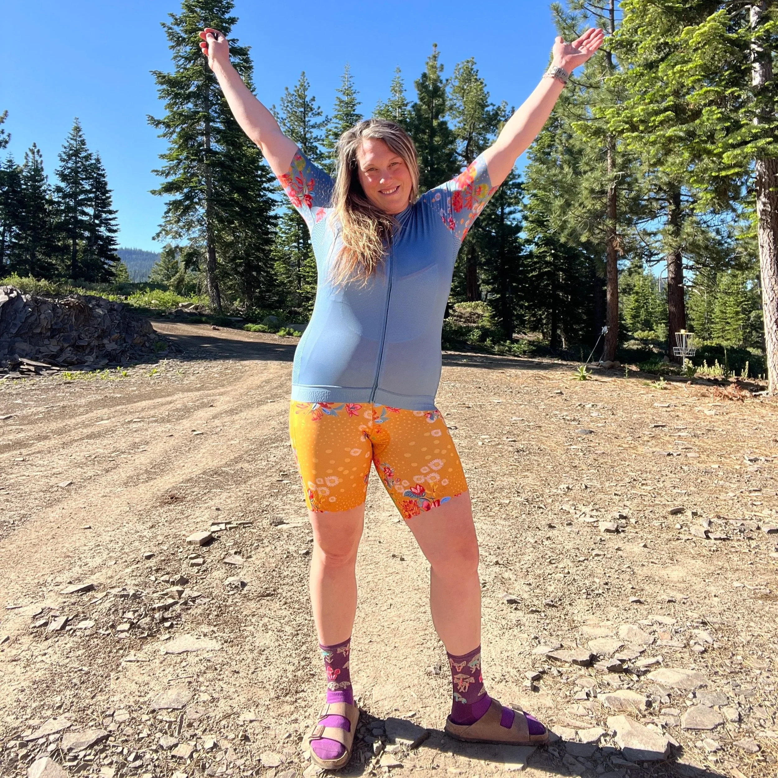 A woman with long wavy hair standing outdoors among tall pine trees, wearing a blue short-sleeve athletic top, orange shorts with floral pattern, purple socks with patterns, and purple sandals, with arms raised in greeting or celebration.