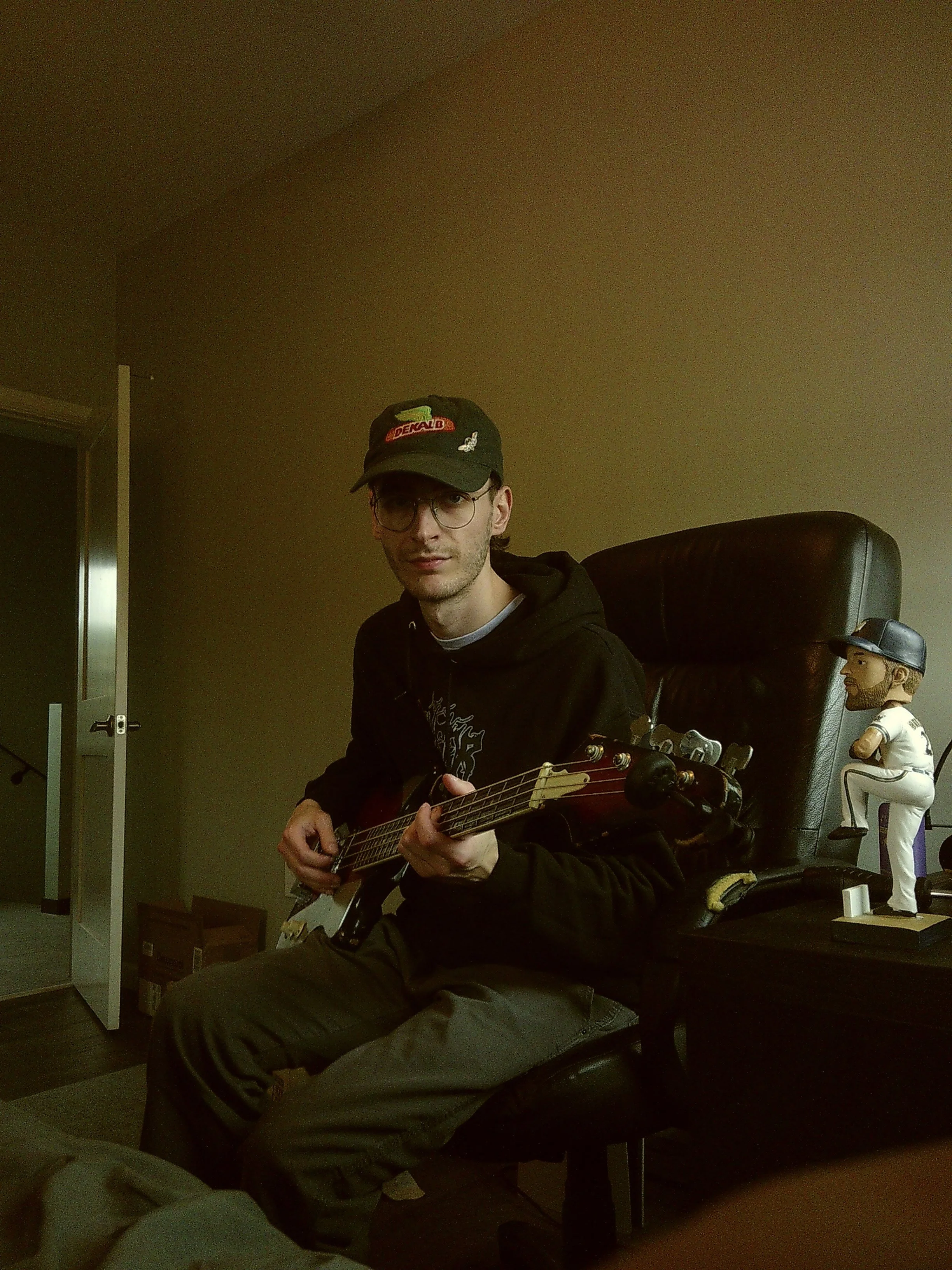 A young man sitting in a black leather office chair, holding an electric guitar, wearing a black hoodie, khaki pants, glasses, and a baseball cap. There is a bobblehead figurine of a baseball player in a white uniform on a table beside him.