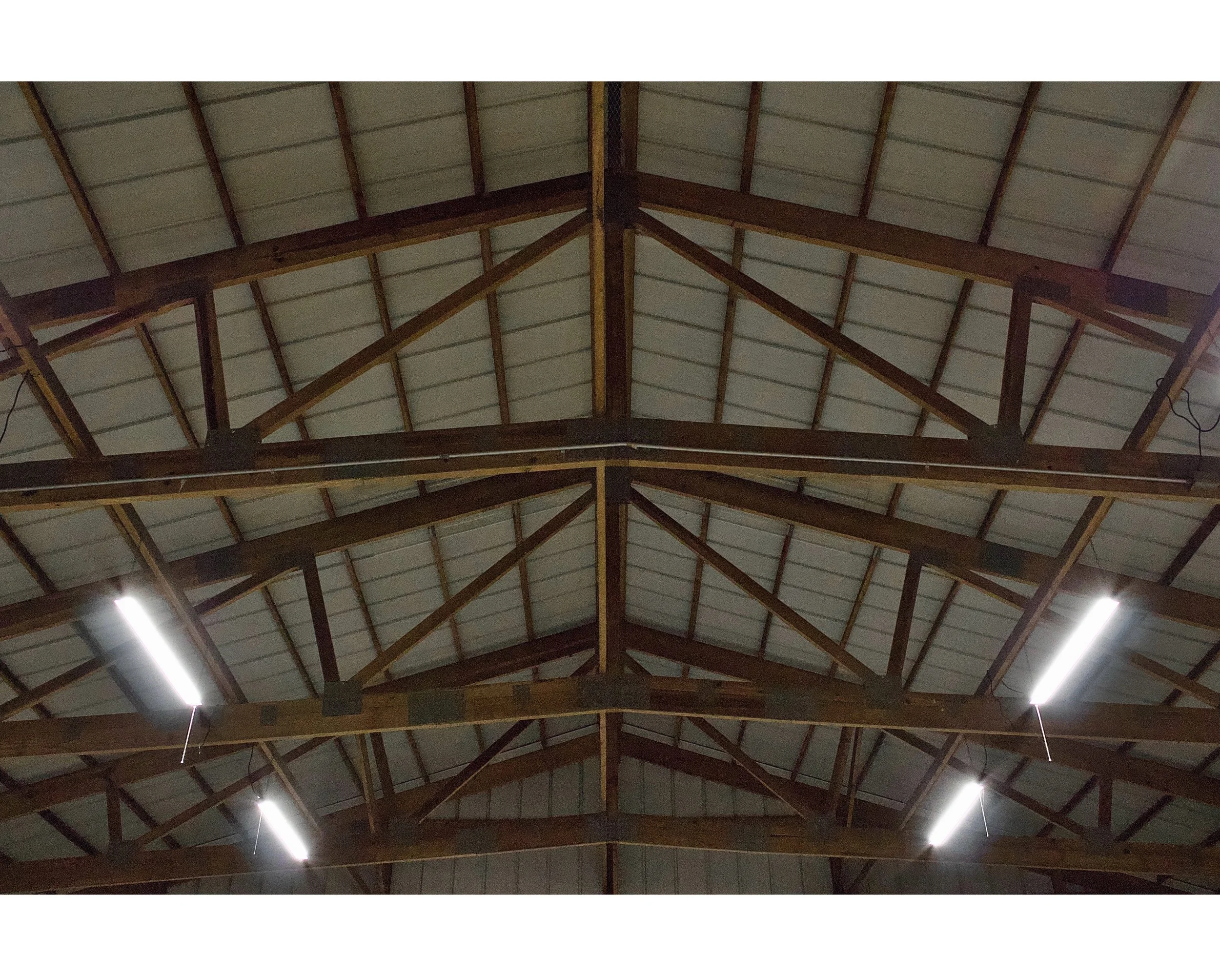 View of a warehouse ceiling with exposed wooden beams and metal roofing panels, illuminated by fluorescent lights.