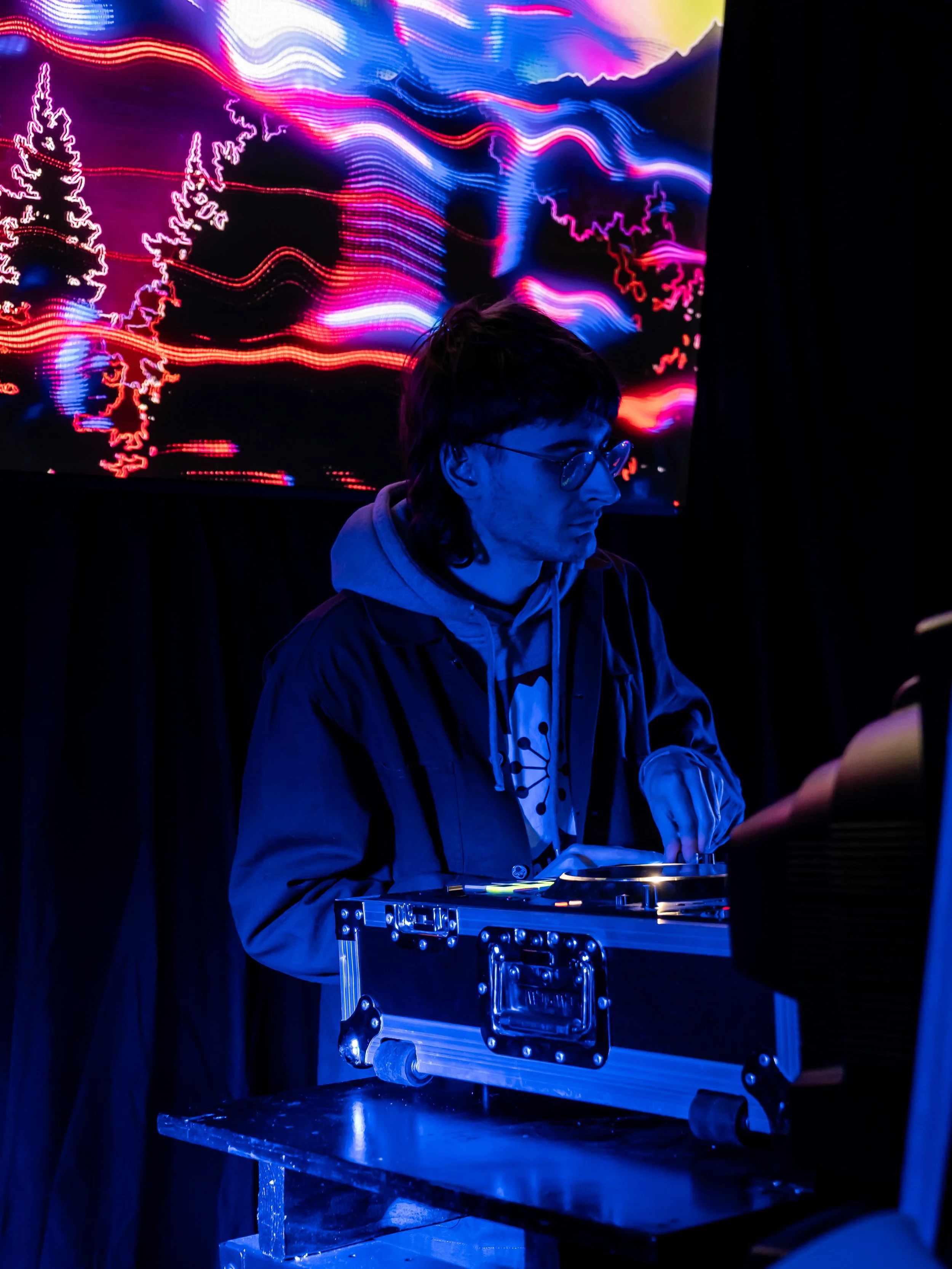 A person with glasses and dark hair is operating DJ equipment at a nightclub or music event. Behind him, vibrant multicolored light patterns are projected onto a screen.