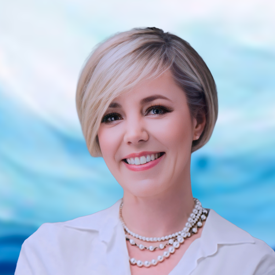 Portrait of a woman with short blonde hair wearing a white shirt and multiple pearl necklaces, smiling at the camera against a gray background.