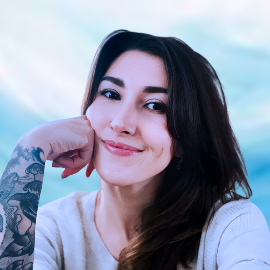 A young woman with dark brown hair, tattoos on her left arm, and a light beige sweater, smiling and resting her chin on her left hand in a relaxed pose.