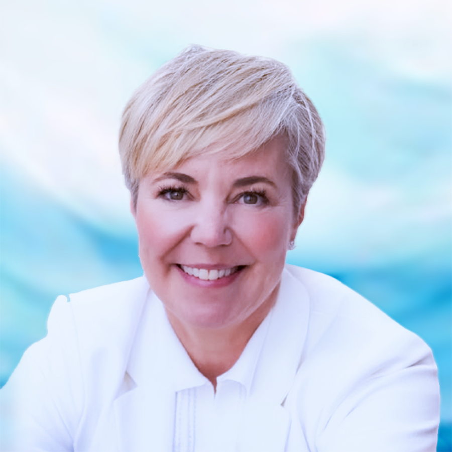 Portrait of a woman with short blonde hair wearing a white shirt and multiple pearl necklaces, smiling at the camera against a gray background.
