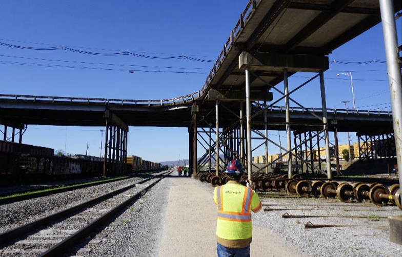 A transportation worker wearing a helmet and high-visibility gear walks next to the railroad tracks that run under the Wilcox Bridge
