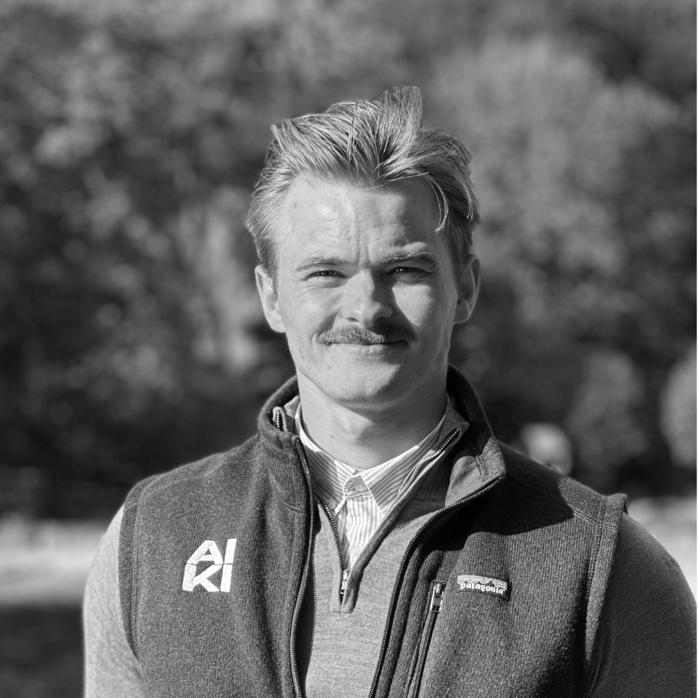 Et portrett av en ung mann med mørkebrunt hår, bart, iført hvit skjorte og svarte slips, stående mot en hvit bakgrunn.