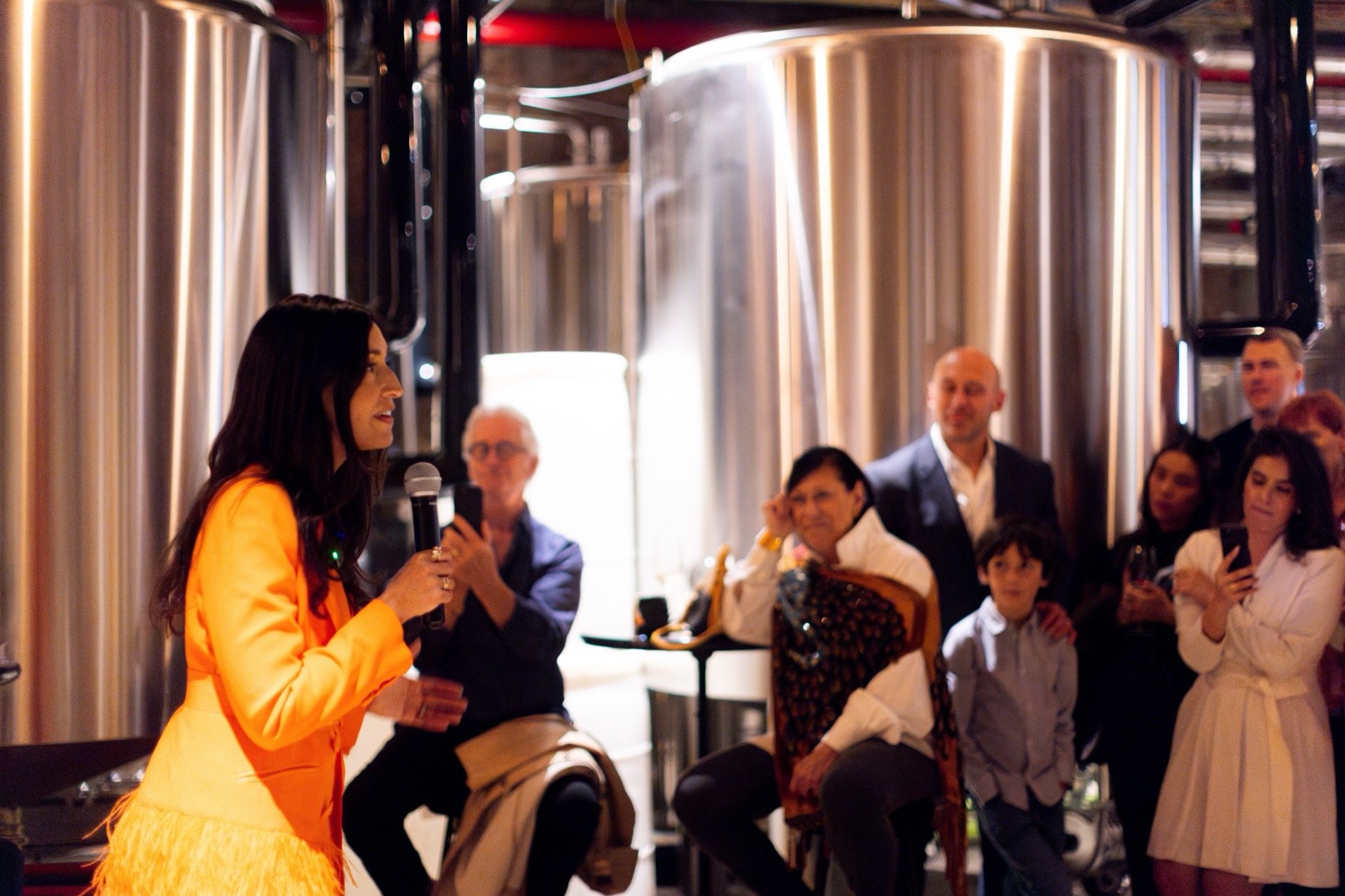 Woman in orange jacket speaking to a crowd in a brewery setting with large metal tanks in the background.