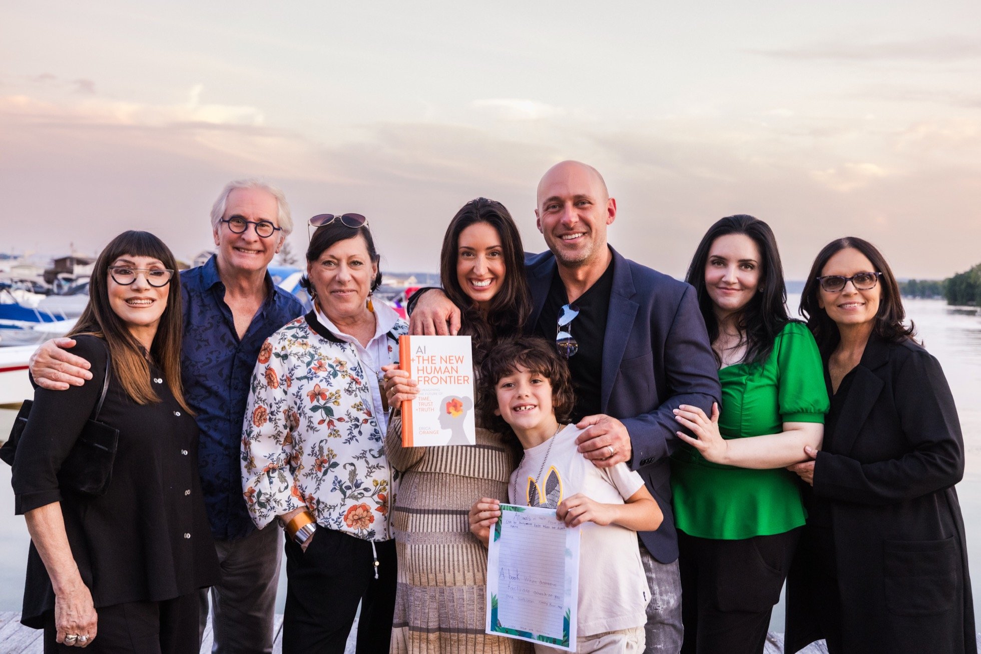 Group of people posing outdoors near water, holding a book titled 'AI: The New Human Frontier.'