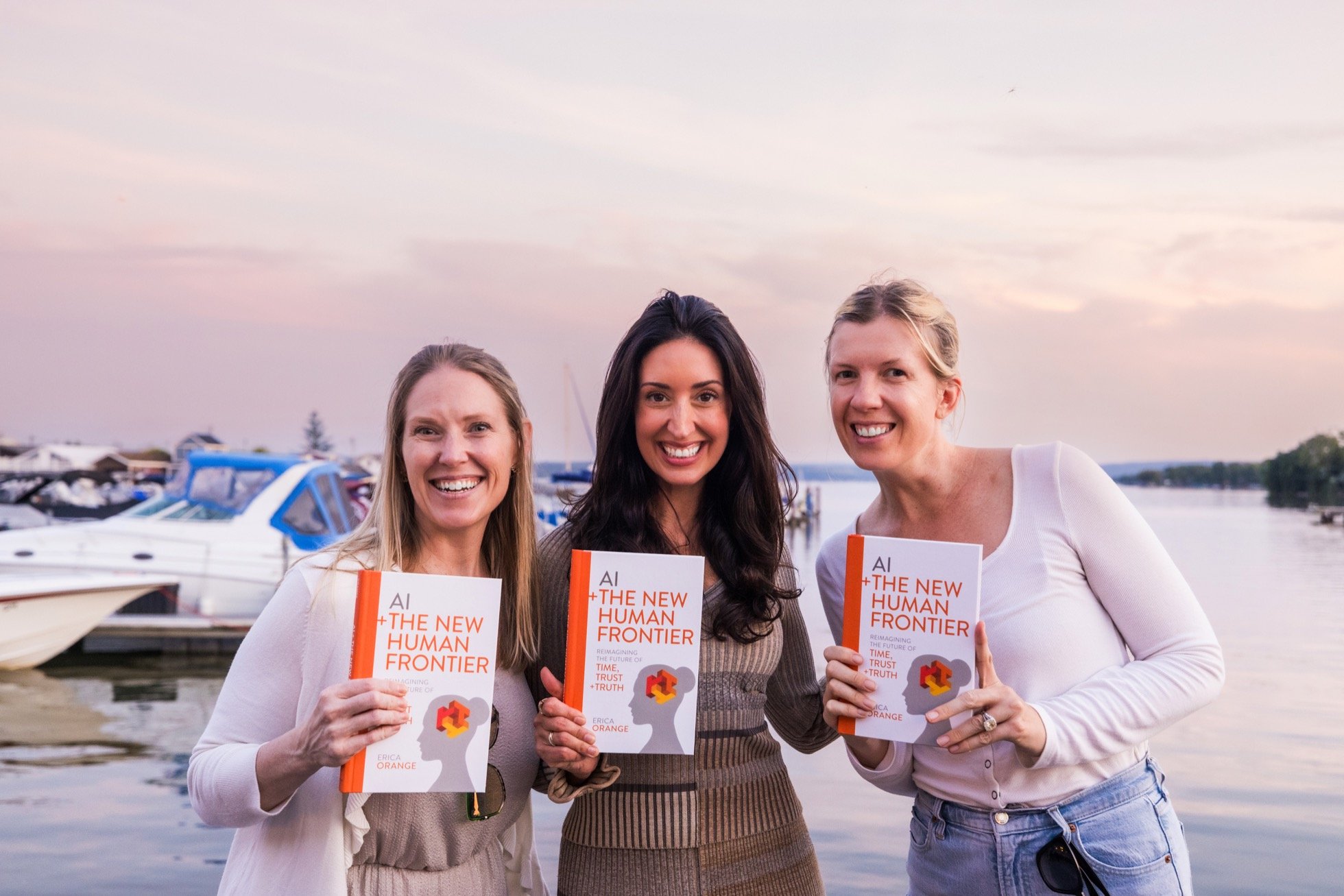 Three women holding a book titled 'AI + The New Human Frontier' outdoors by the water with boats in the background at sunset.