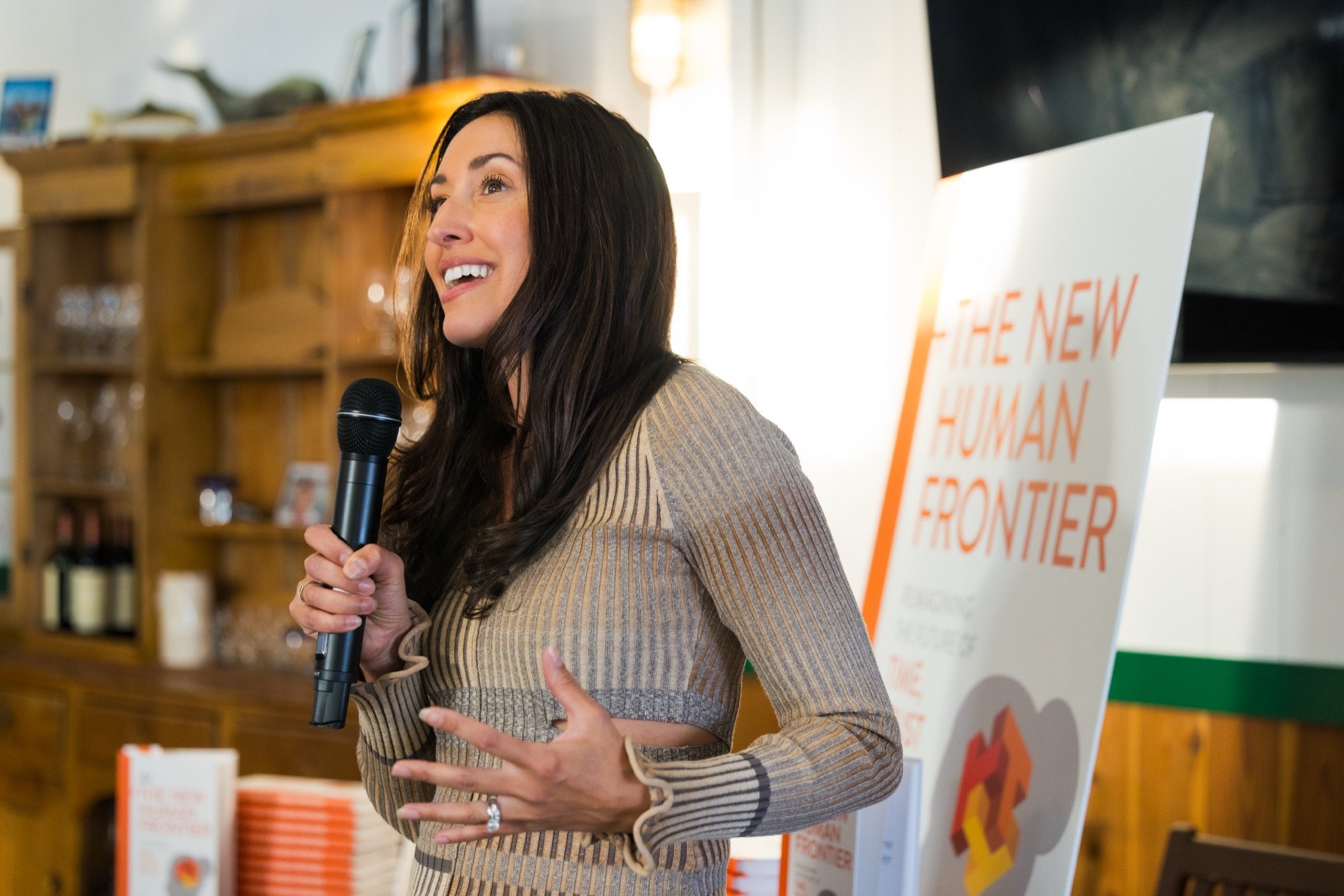 Woman speaking into a microphone at an event, with a display board titled "The New Human Frontier" in the background.