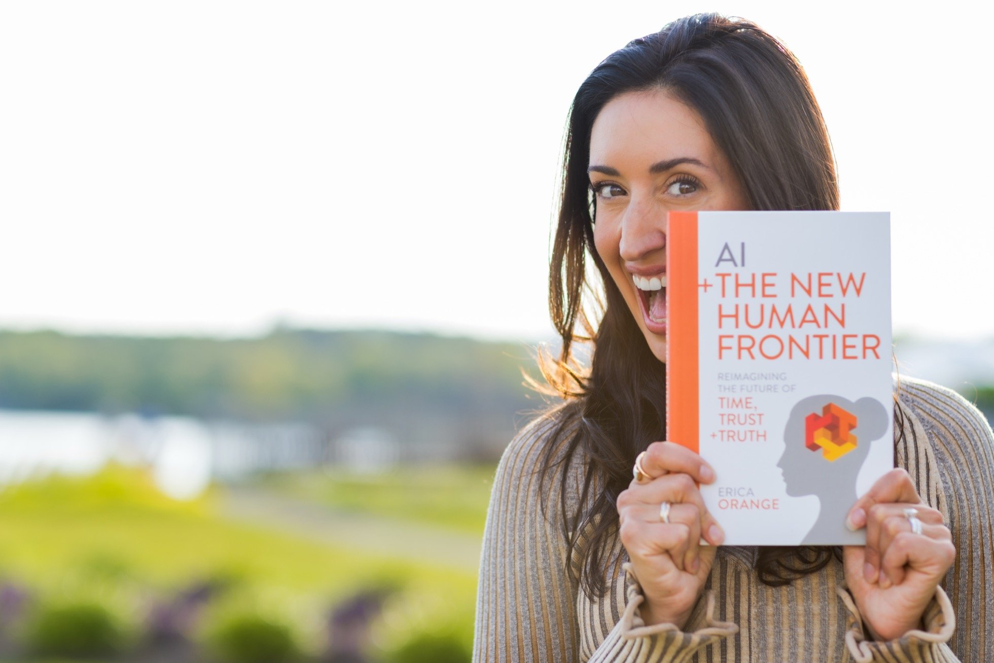 A woman smiling and holding up a book titled 'AI + The New Human Frontier' by Erica Orange, with a blurred outdoor background.