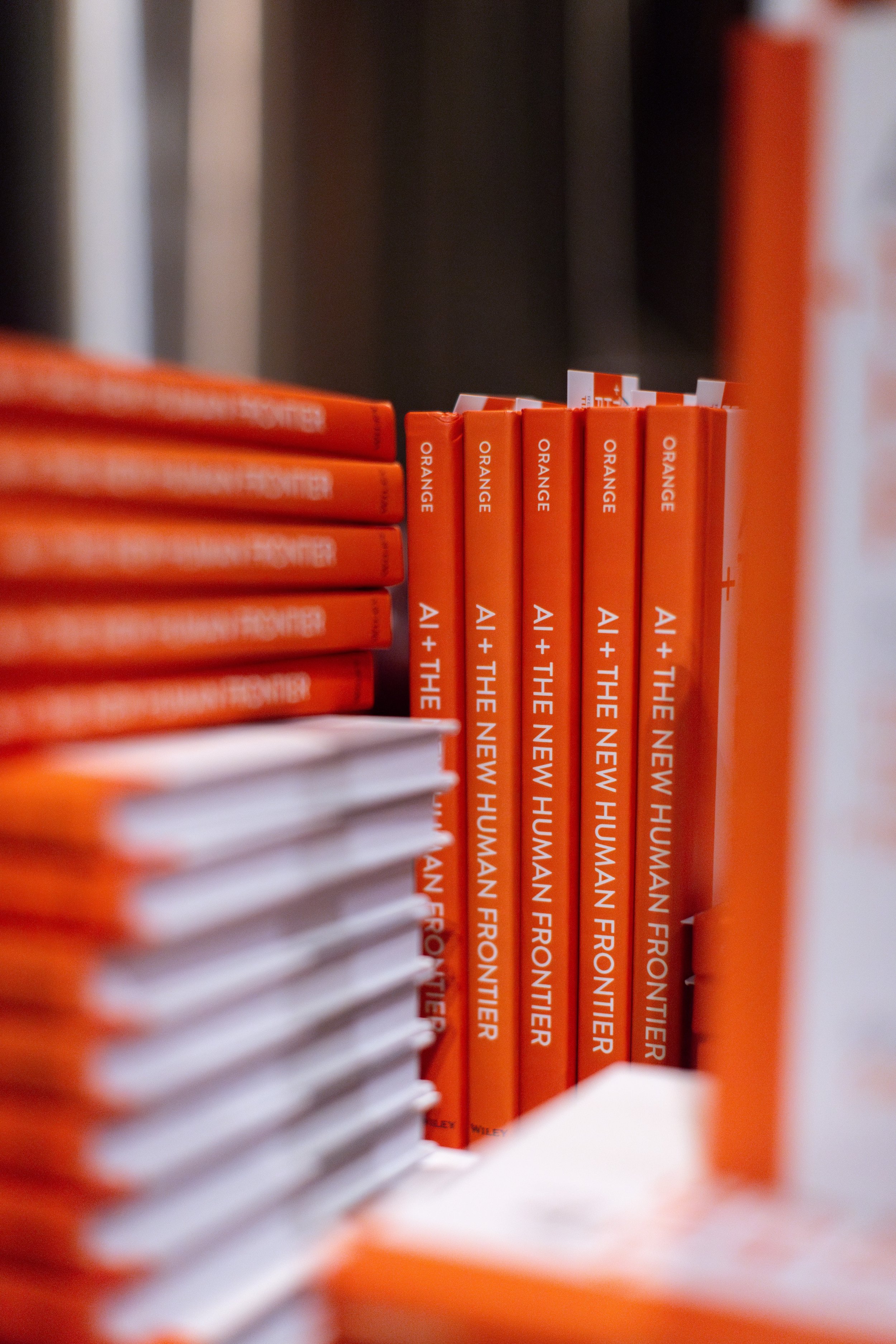 Stacks of orange books titled "AI + The New Human Frontier" in a bookstore or display.