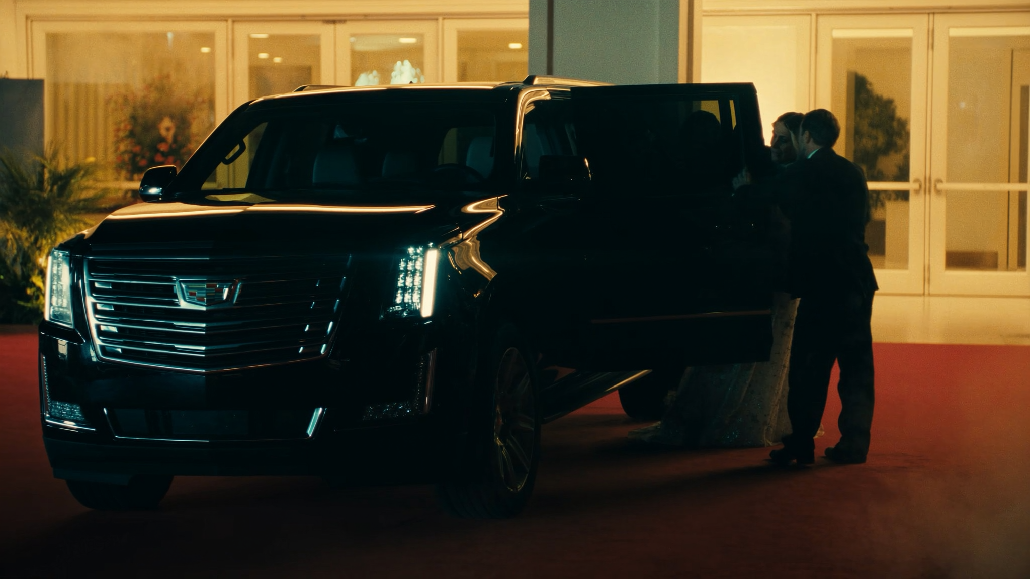 A black Cadillac Escalade limousine parked outside a building at night with two men in suits standing next to it, one opening the door.