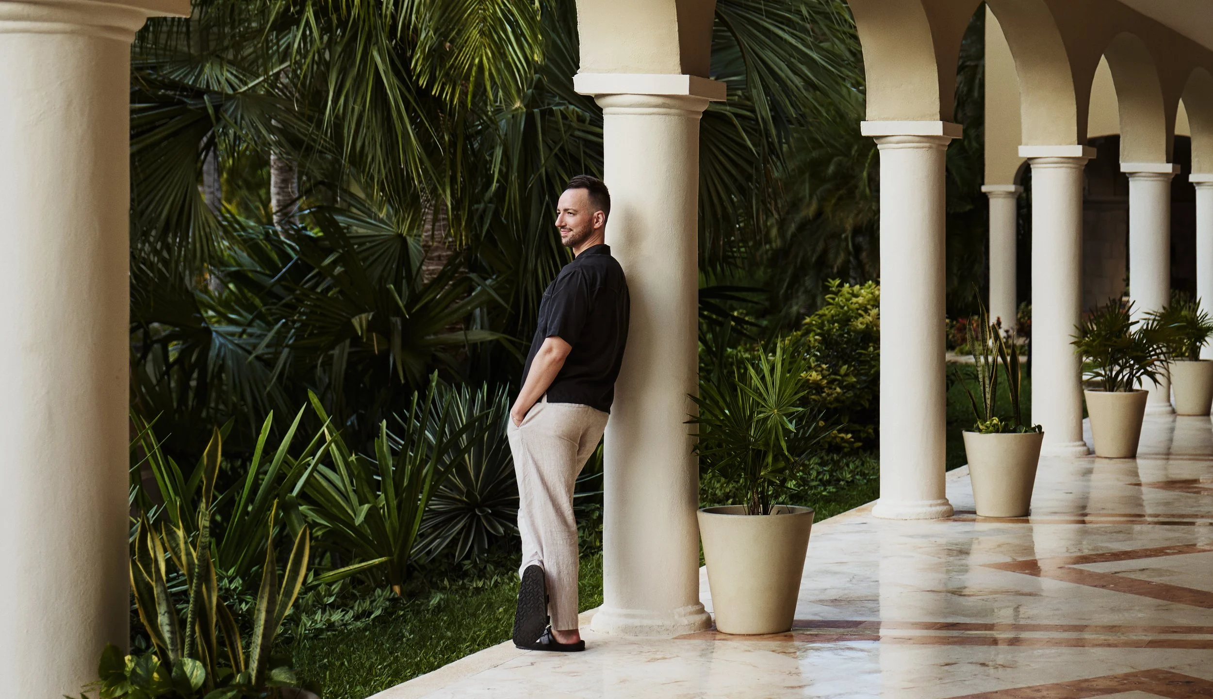A man leaning against a column on a tiled patio, surrounded by potted plants and lush greenery.