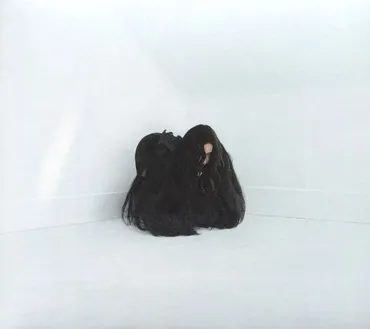 A person with long black hair sitting on the floor against a white wall, their face partially obscured. Album art.