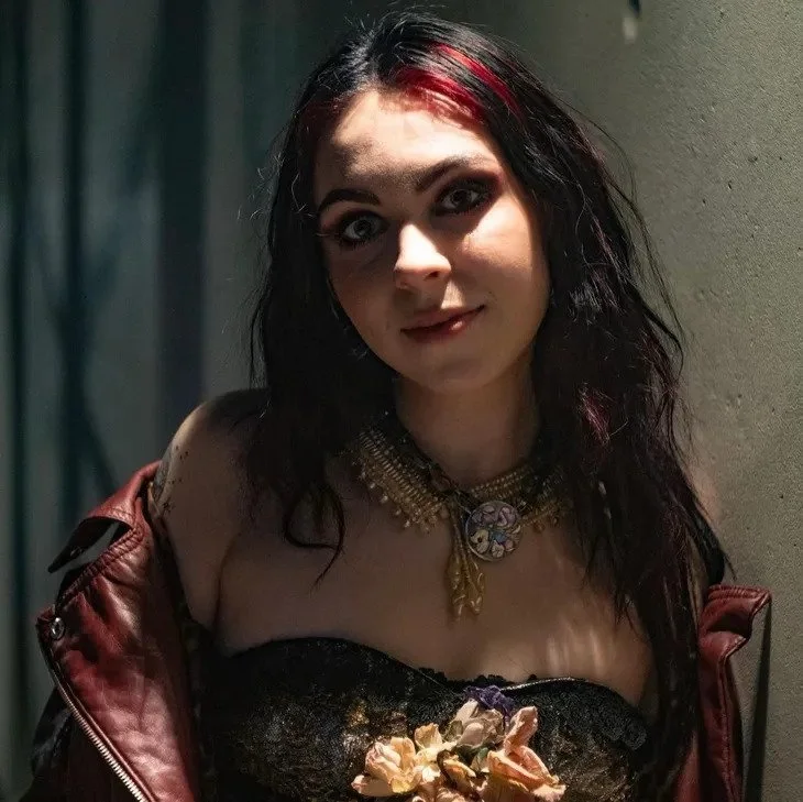 Young woman with black and red hair, wearing a black strapless top, a gold necklace with a large pendant, and a red leather jacket, standing against a wall.