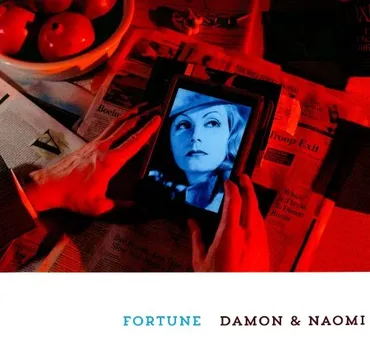 Person holding a smartphone with a photo of a woman on the screen, on a table covered with newspapers and apples, illuminated by red and blue lighting. Album art for "Fortune" by Damon and Naomi