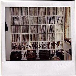 Wall with shelves filled with vinyl records and a sign that says 'LCD SOUND SYSTEM'. Album artwork.