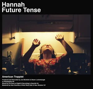 A person standing in a kitchen with arms raised and head tilted back, illuminated from above, with the text 'Hannah Future Tense' and 'American Trappist' overlaid. Album Cover.