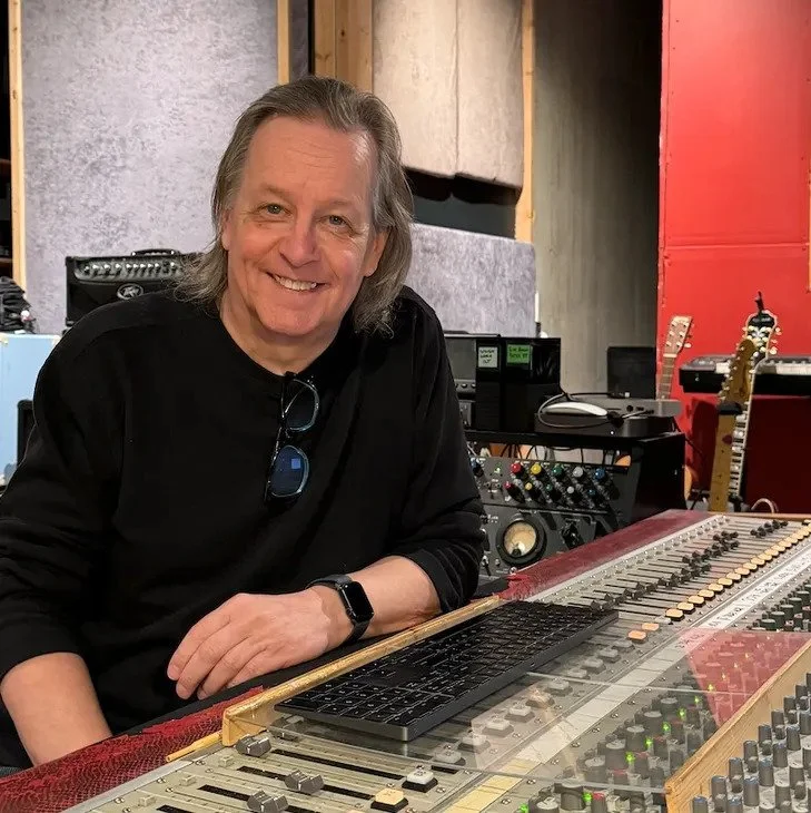 Smiling man with long gray hair sitting at a large audio mixing console in a recording studio.