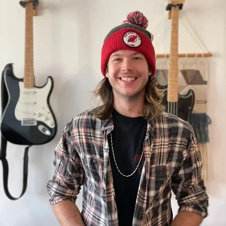 Young man with long hair wearing a red and gray knit beanie, plaid shirt, and black t-shirt, smiling in a room with guitars hanging on the wall.