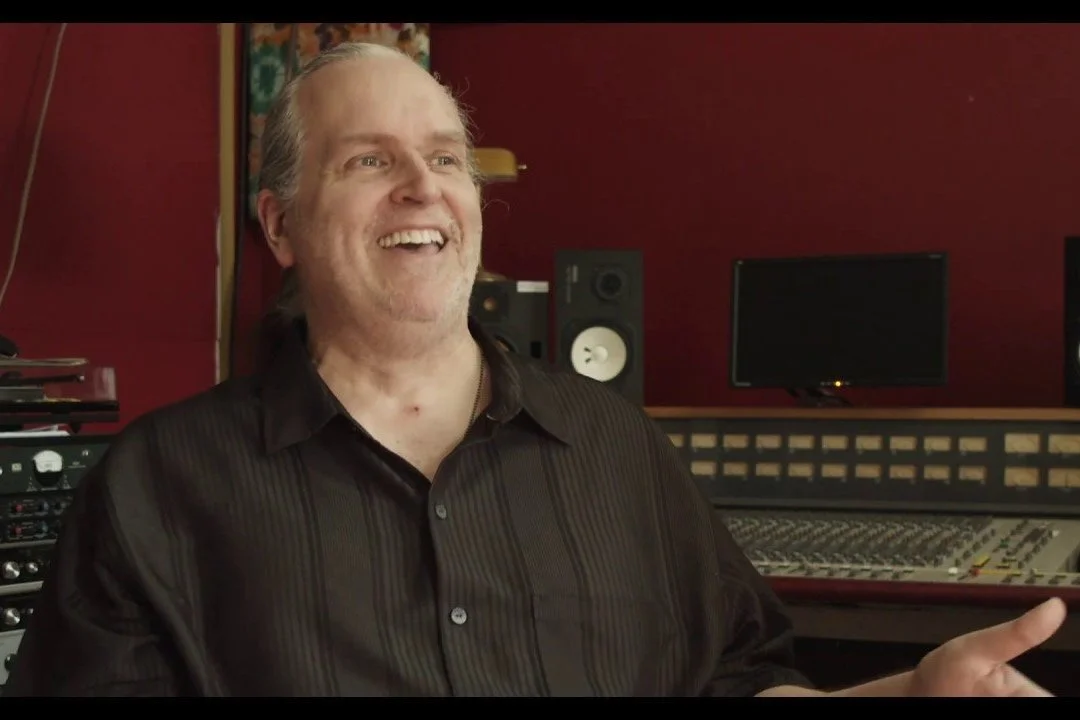 Man in black (Alan Douches) shirt smiling in recording studio with audio equipment and monitors in the background.