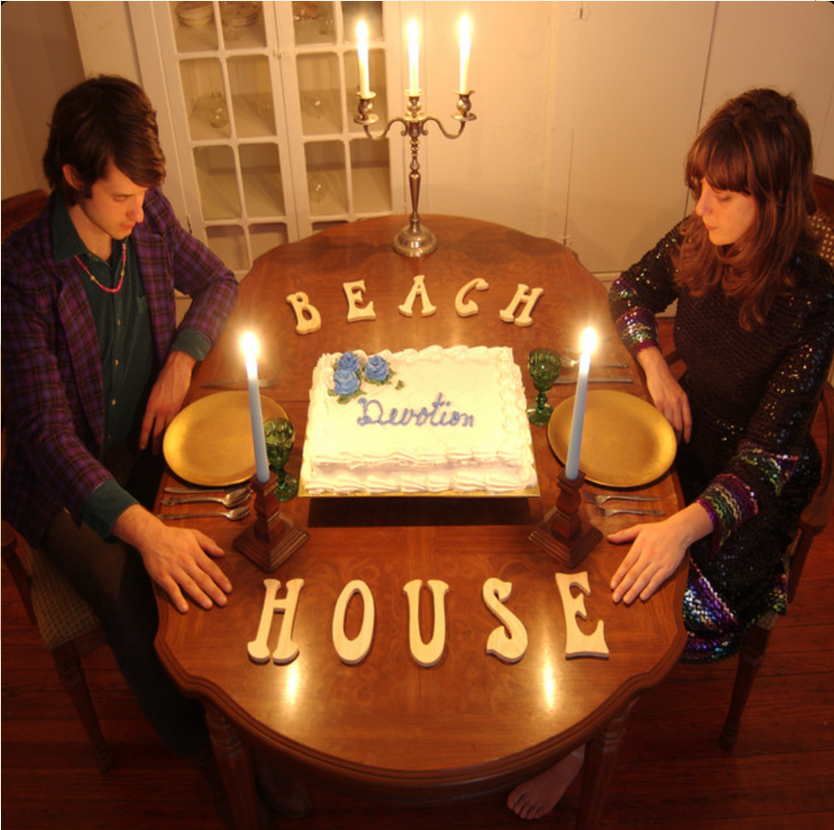 A man and woman sit at a dinner table with a birthday cake and lit candles, surrounded by letter blocks spelling 'BEACH' and 'HOUSE', in a cozy room with a candle center piece. Album cover for 'Devotion.'