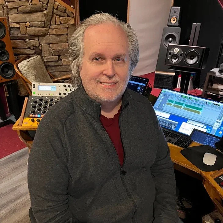 A man with gray hair and a beard smiling in a music studio with speakers, sound equipment, and a computer displaying audio editing software.