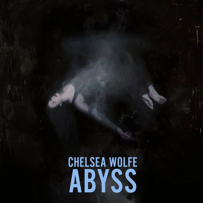 A woman lying on a dark surface with her hair spread out, wearing a black dress, with smoke or mist surrounding her, and the text 'Chelsea Wolfe Abyss' below. Album artwork.