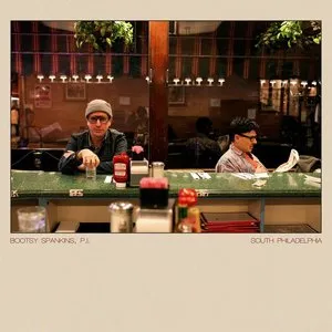 Two men sitting at a bar counter, one in a beanie and sunglasses, the other reading a newspaper, with a window and hanging plants in the background. Album cover.