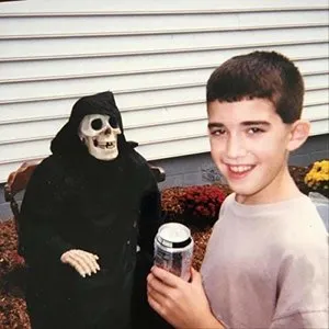 A young boy smiling and holding a beverage next to a skeleton figure dressed in dark clothing.