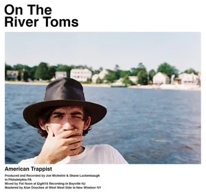 A young man with a hat covering part of his face, standing near a river with houses on the opposite bank in the background, titled 'On The River Toms'. Album cover.