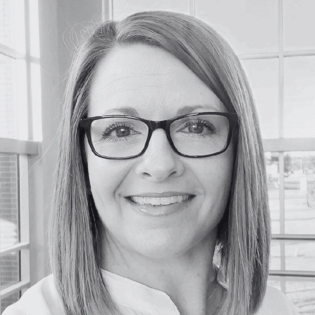 A black and white close-up portrait of a smiling person wearing glasses, standing in front of a window with a grid-like pattern.
