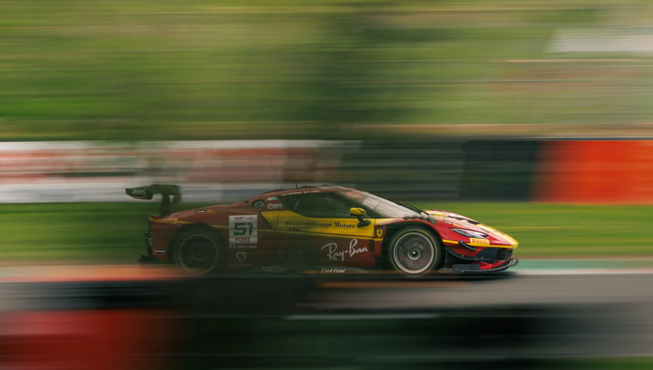 A red Ferrari race car with yellow accents speeding on a racetrack, with motion blur in the background.