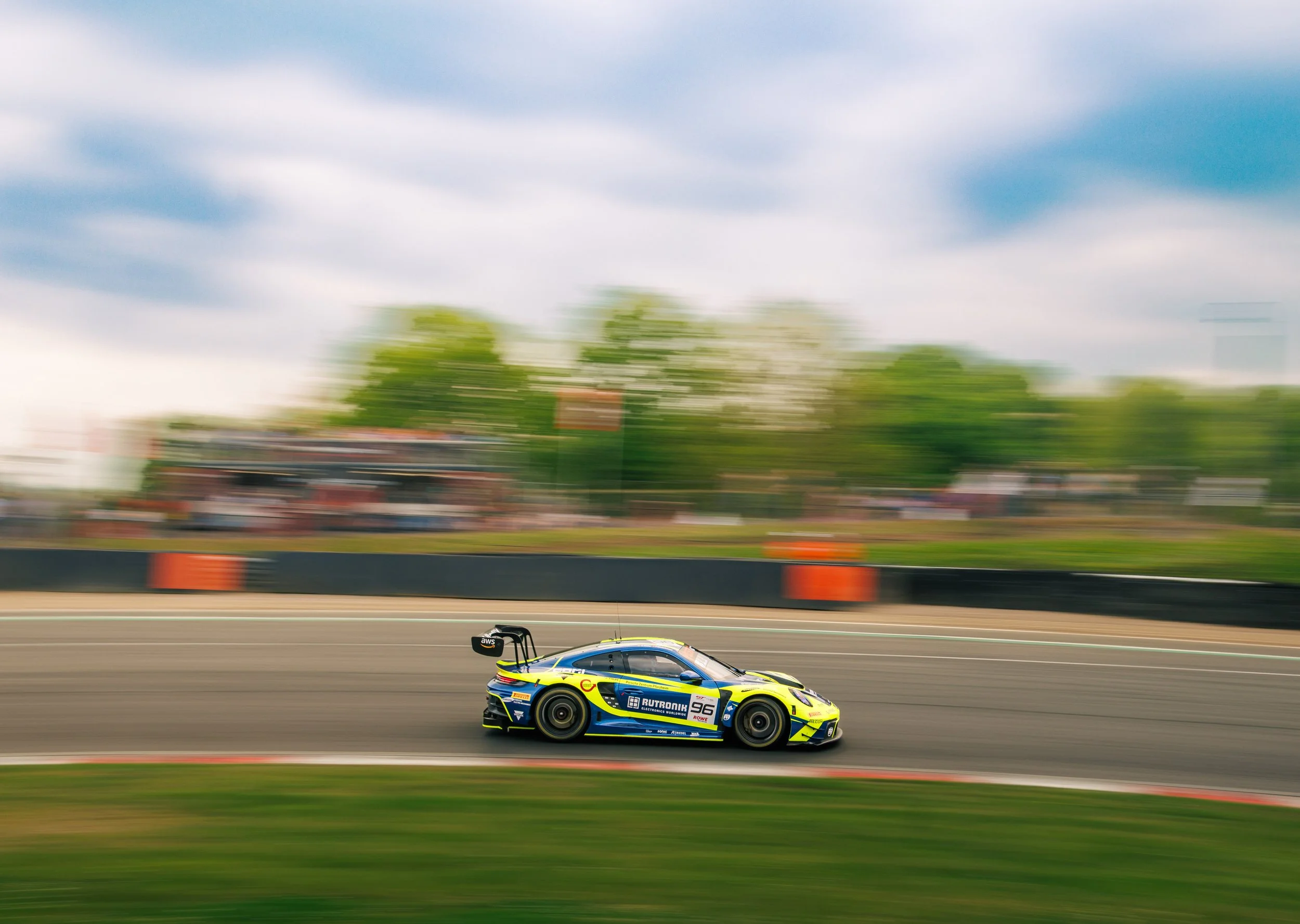 A racing car speeding on a race track with motion blur, surrounded by green trees and a cloudy sky.