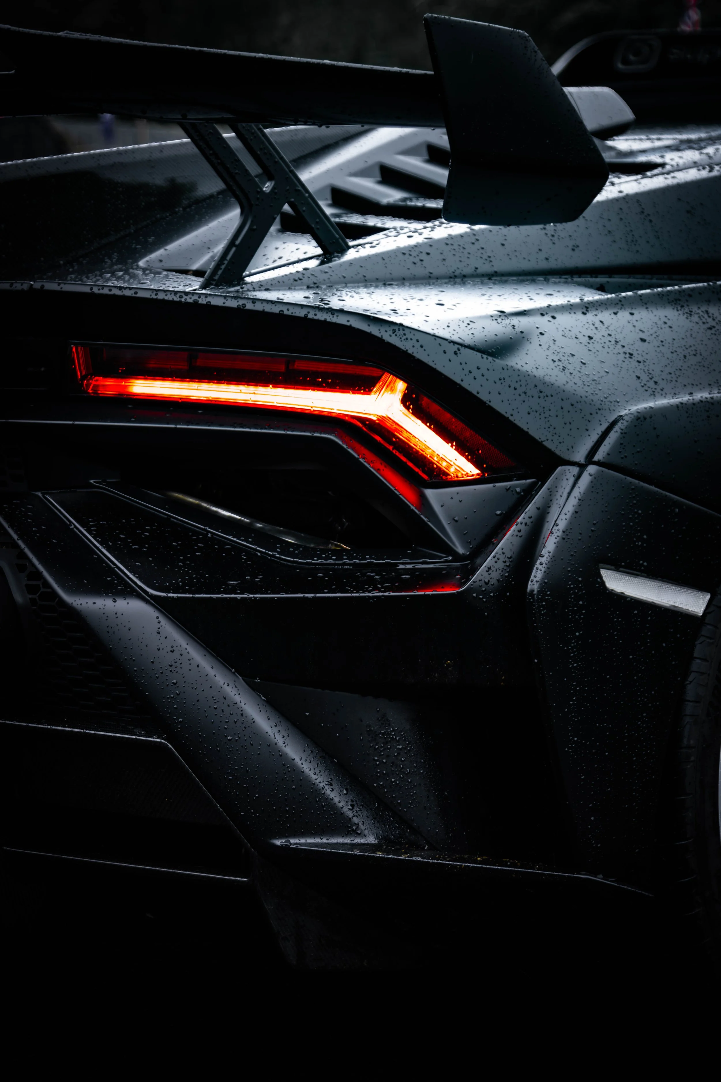 Close-up of the rear corner of a black sports car with glowing red tail light, rain droplets on the surface, and a large rear spoiler.
