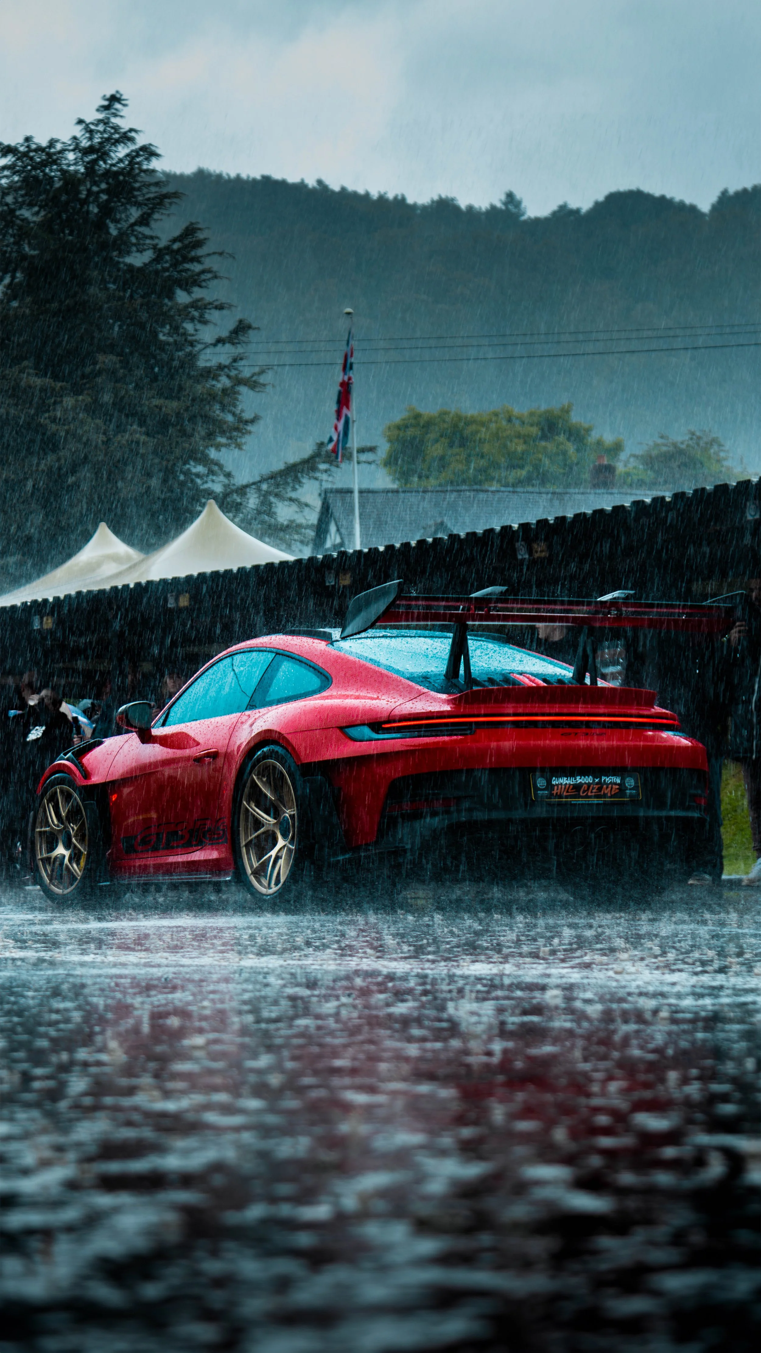 Red sports car, likely a Porsche, parked in heavy rain with mountains and trees in the background. The scene appears to be at a racing event or gathering.