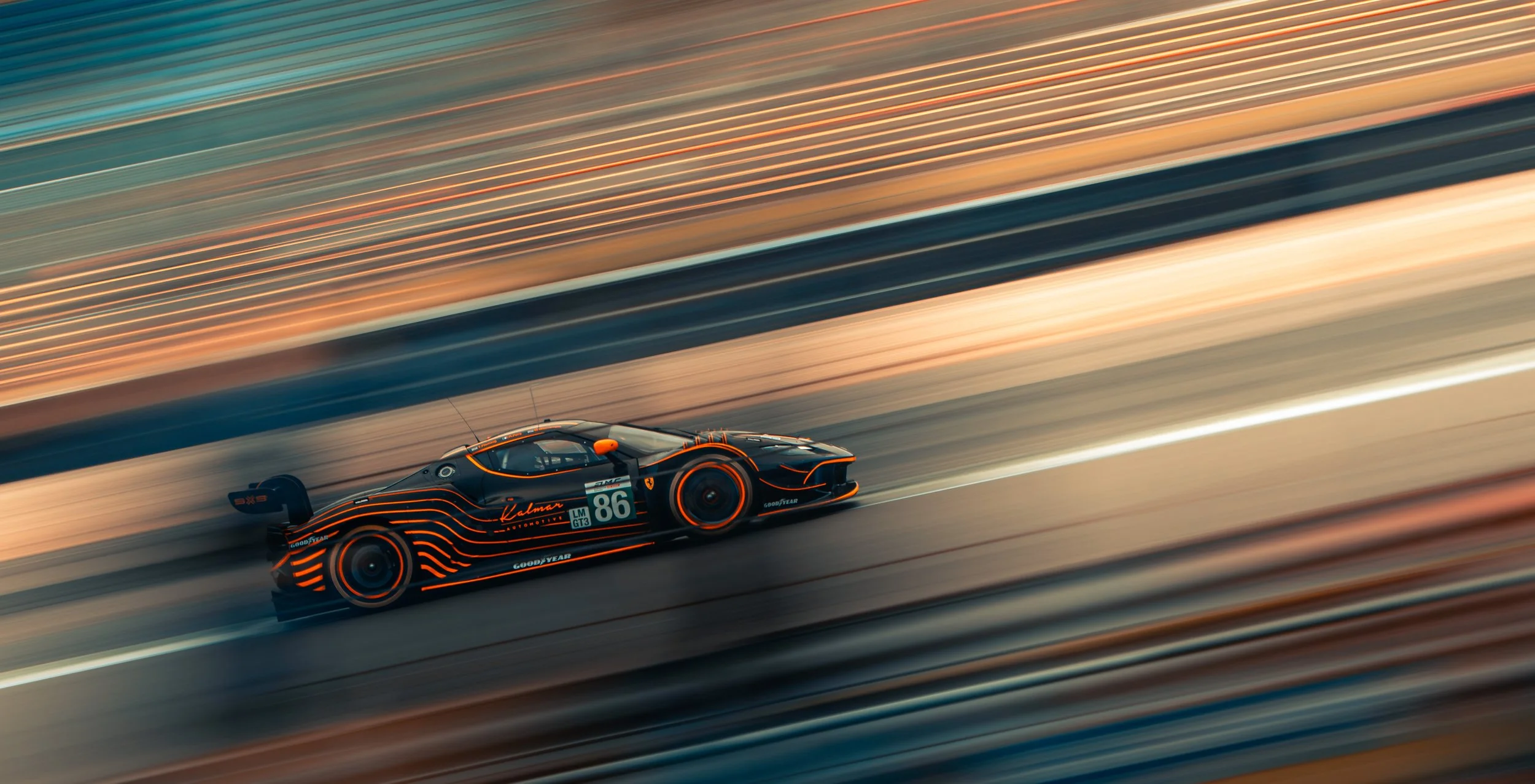A black race car with orange accents and stripes speeding on a racetrack during sunset, creating a motion blur effect.