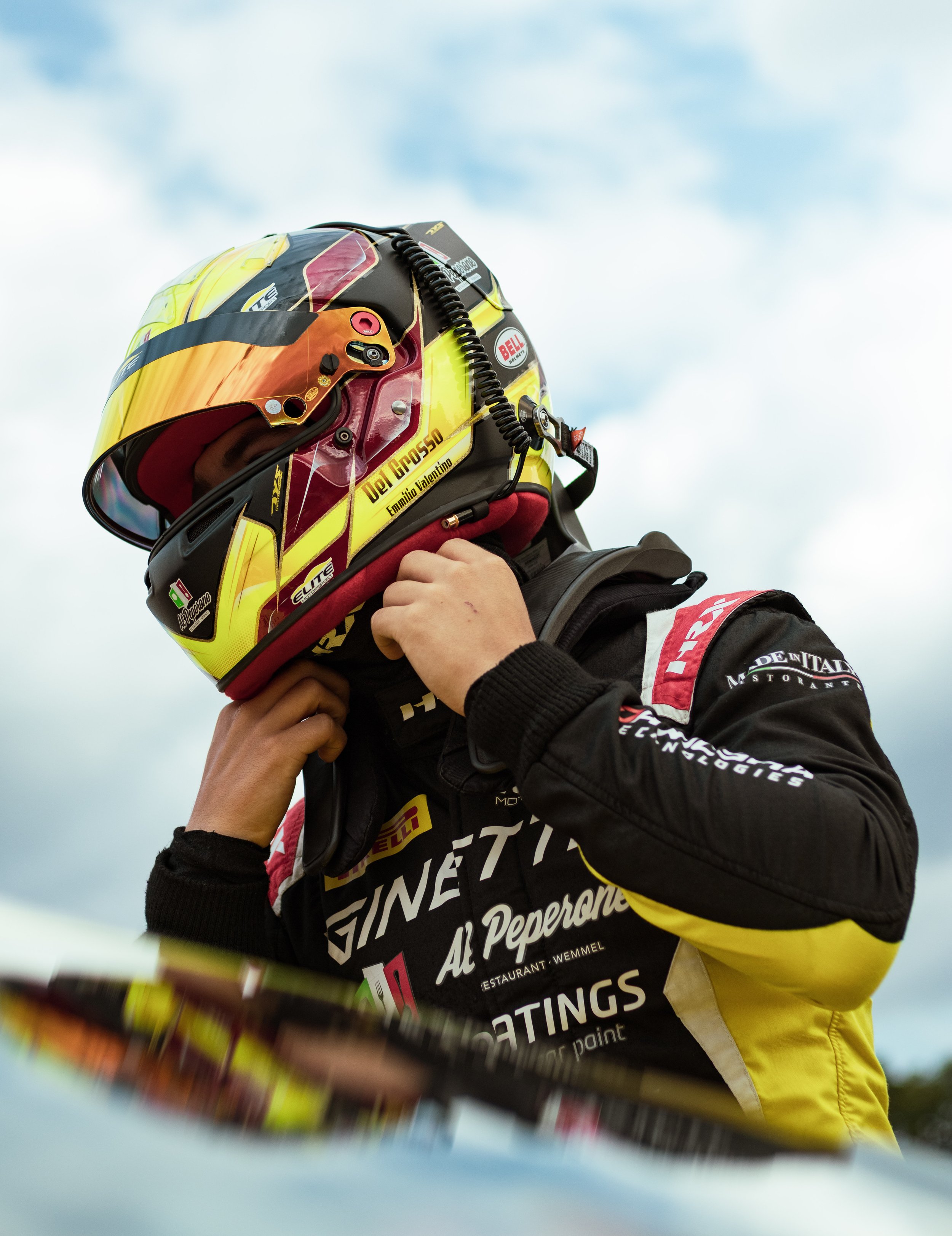 Race car driver adjusting helmet strap before a race, wearing a racing suit with various sponsor logos.