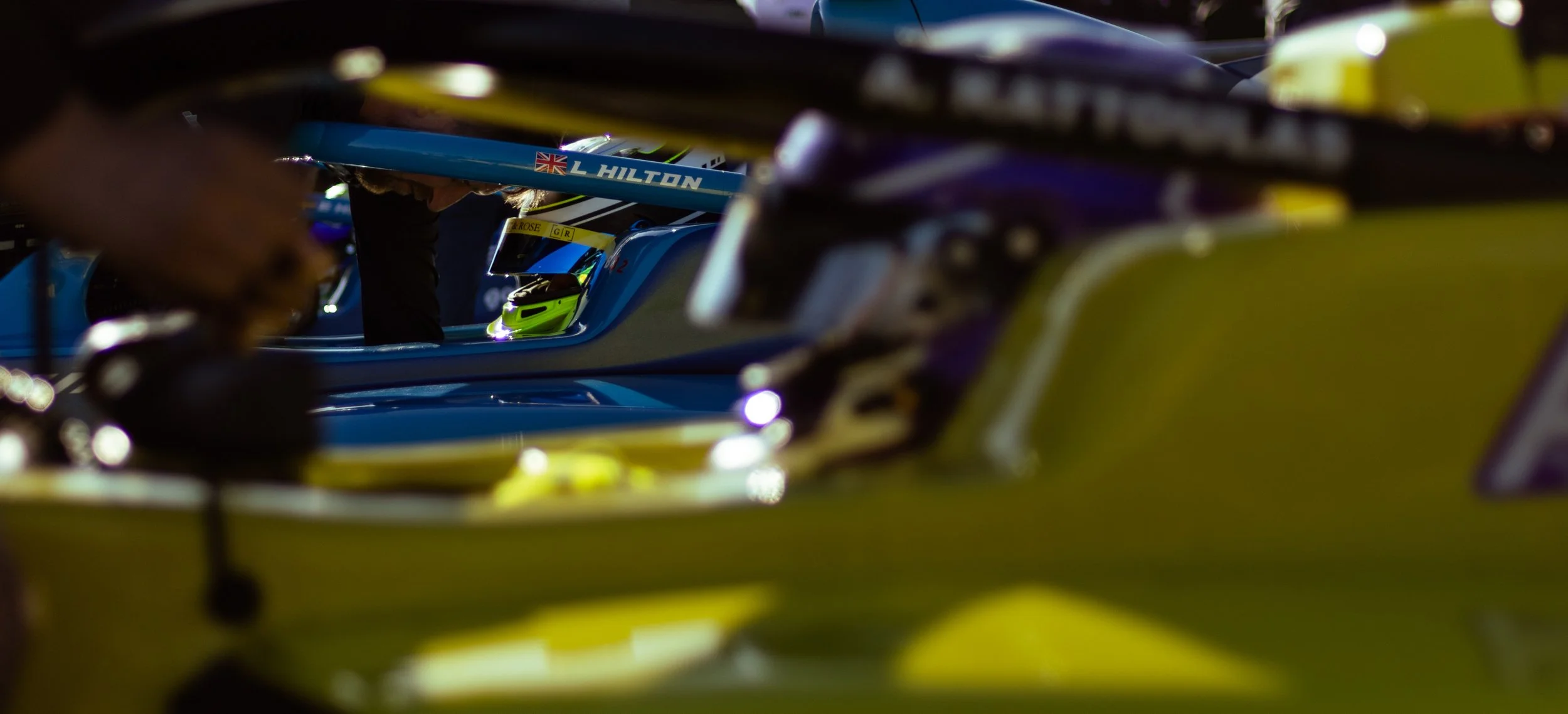 Close-up of Formula 1 car in pit lane with a driver wearing a helmet, featuring sponsorship and branding, focus on cockpit area with team members working around it.