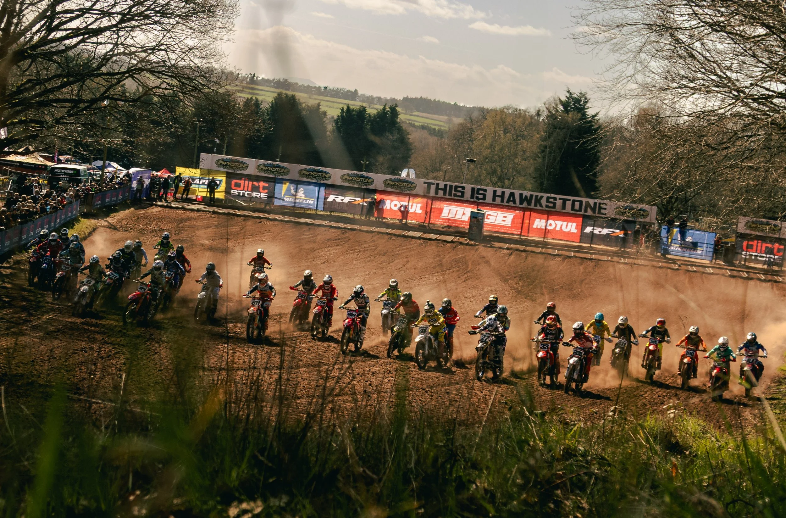 Motocross race on a dirt track with riders kicking up dust, spectators along the sides, and banners displaying sponsors and 'This is Hawkstone' in the background.