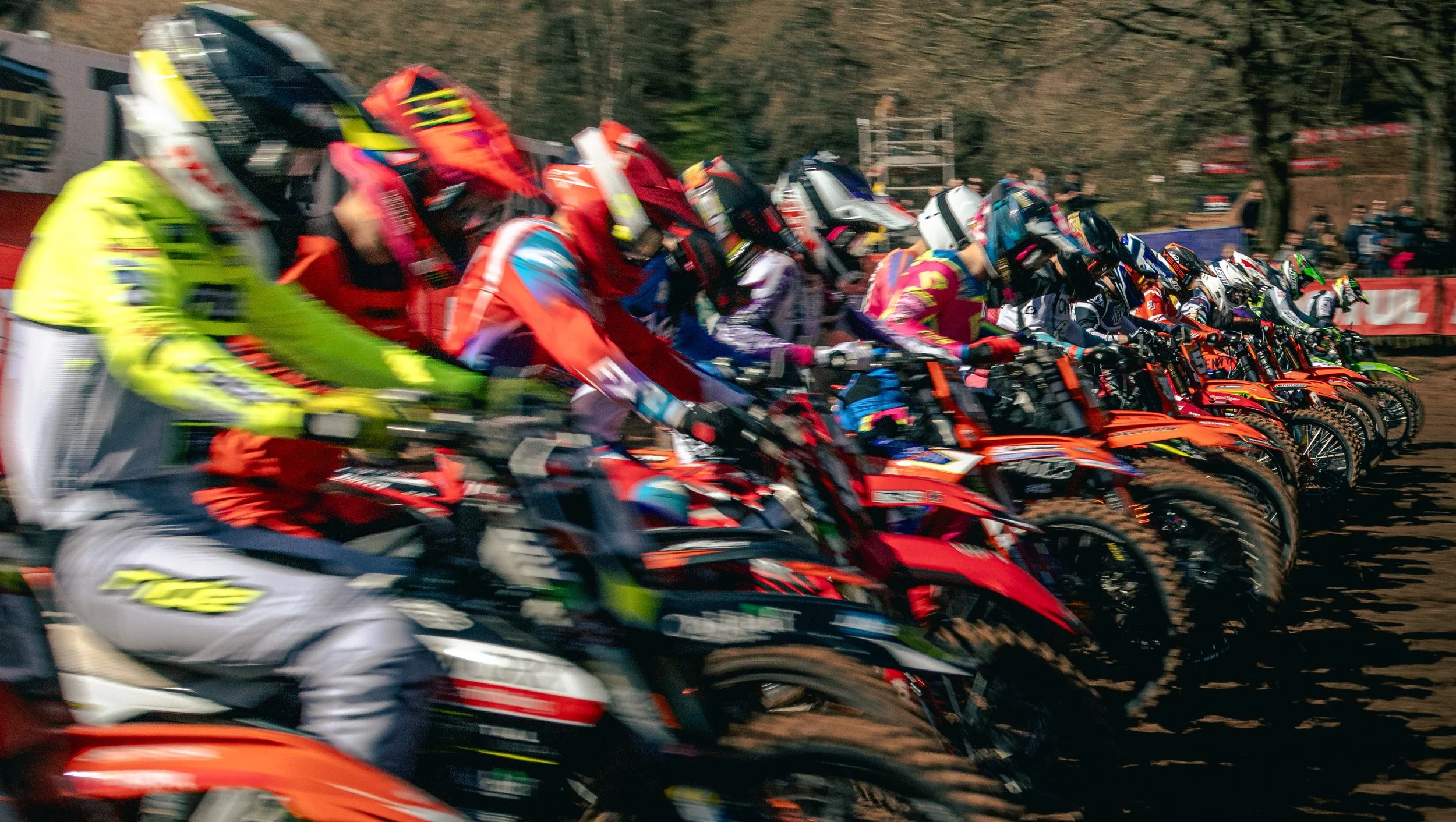 Motocross race with multiple riders wearing colorful gear and helmets, lined up on dirt bikes at the start of the race, with motion blur indicating high speed.
